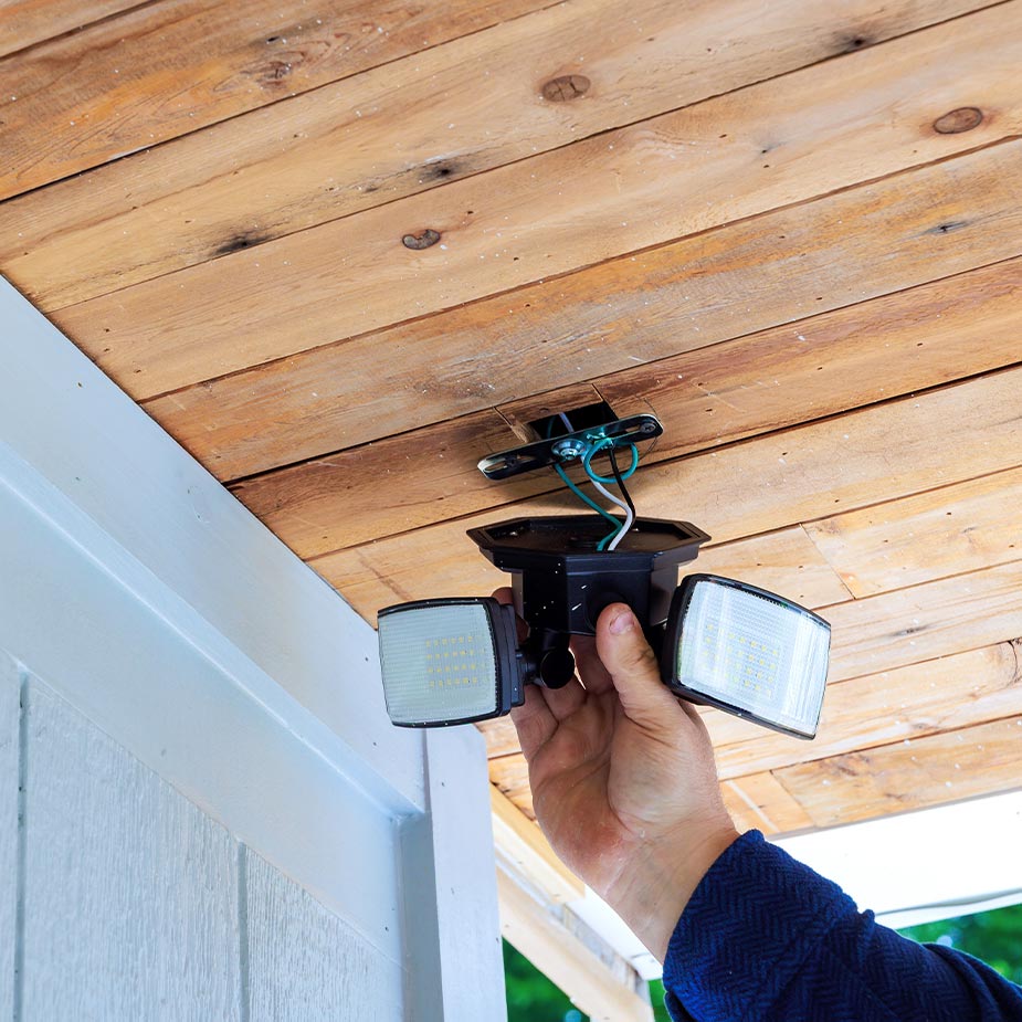 Person installing a two-head outdoor LED floodlight on a wooden porch ceiling.