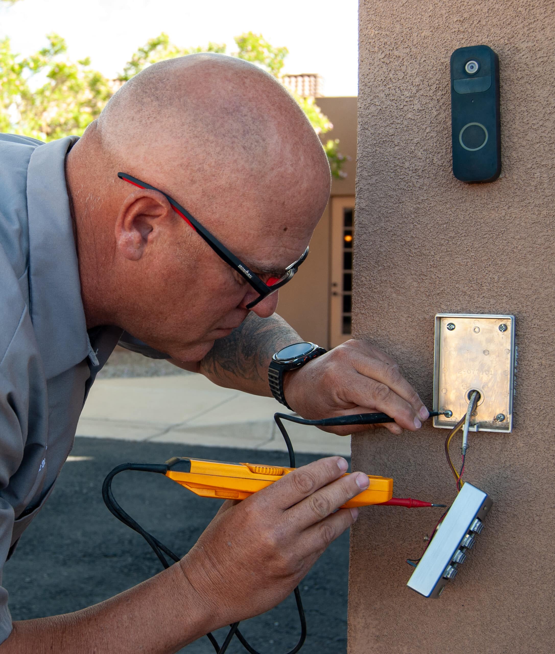 Man with a shaved head wearing glasses uses a handheld tester on an exterior doorbell/intercom panel, wires exposed on a stucco wall nearby.