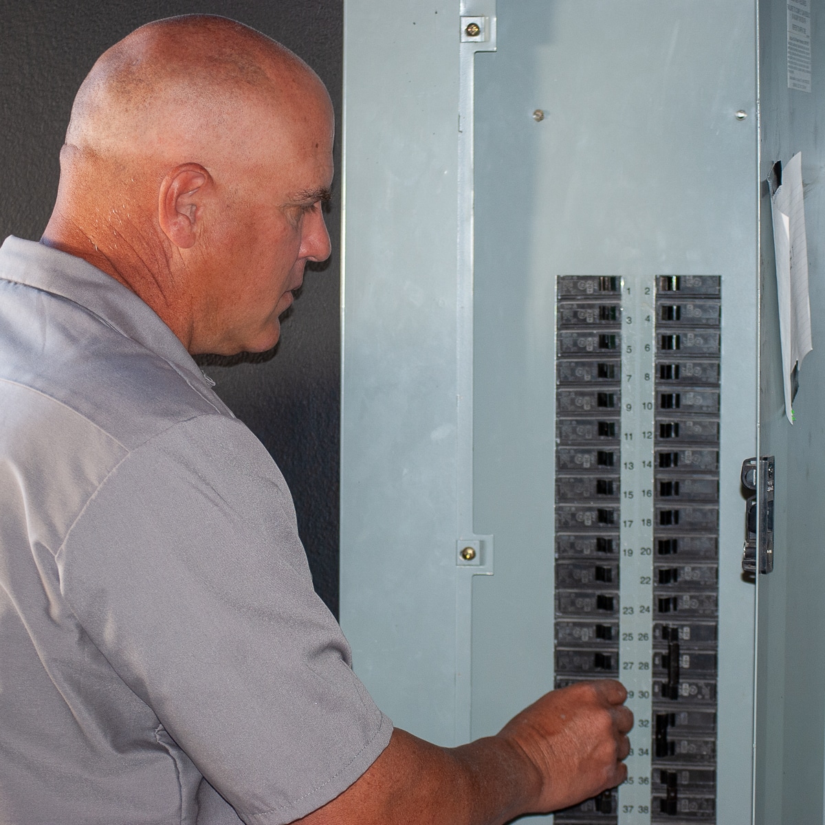 Man in a gray shirt examining or adjusting a vertical row of circuit breakers in an electrical panel.