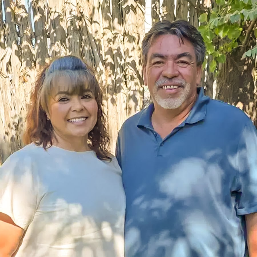 Smiling couple standing outdoors with trees in the background, woman in a light top and man in a blue polo shirt.