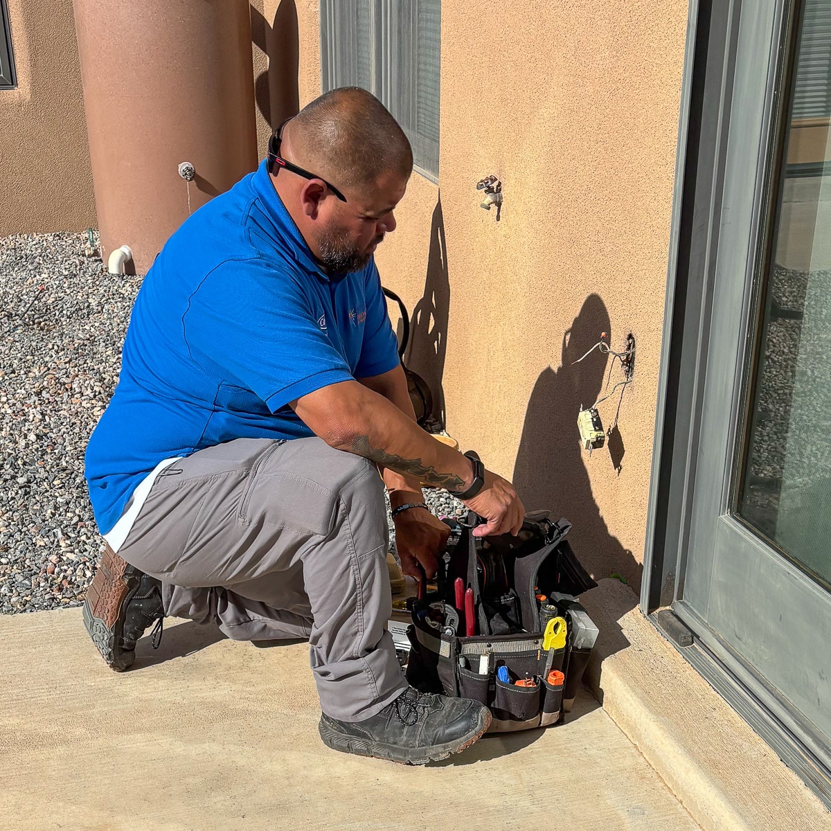 Electrician kneeling on a concrete patio beside a house exterior, working with a tool bag full of pliers and wires near an outdoor outlet.