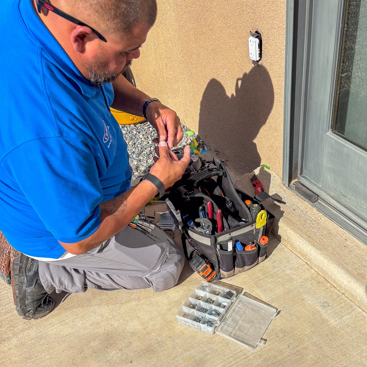 A technician kneels outside a building, sorting small electrical parts from a tool bag on the ground.