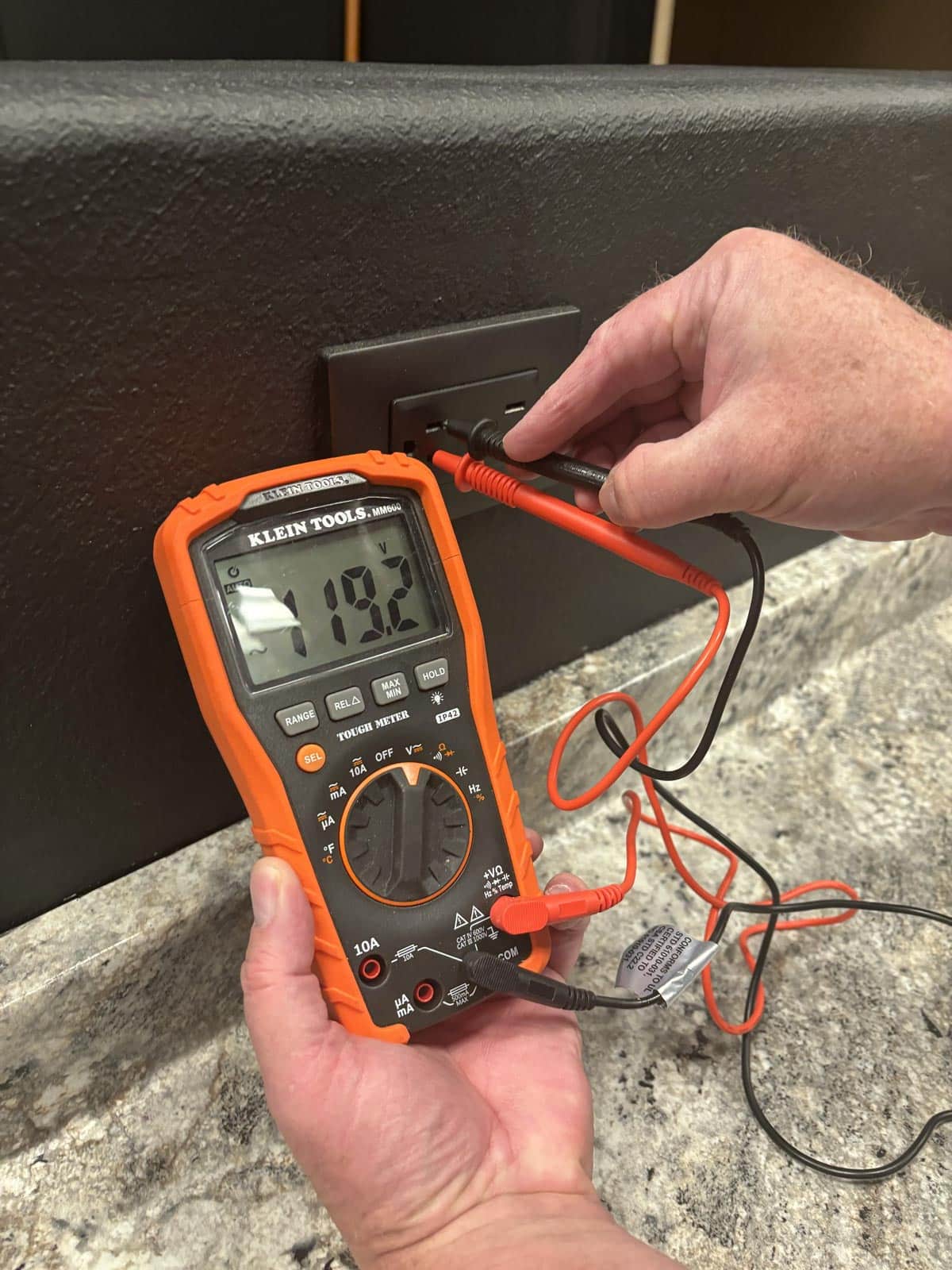 Person using a digital multimeter to test the voltage of a wall outlet, red probes plugged into the socket; display shows about 120 V.